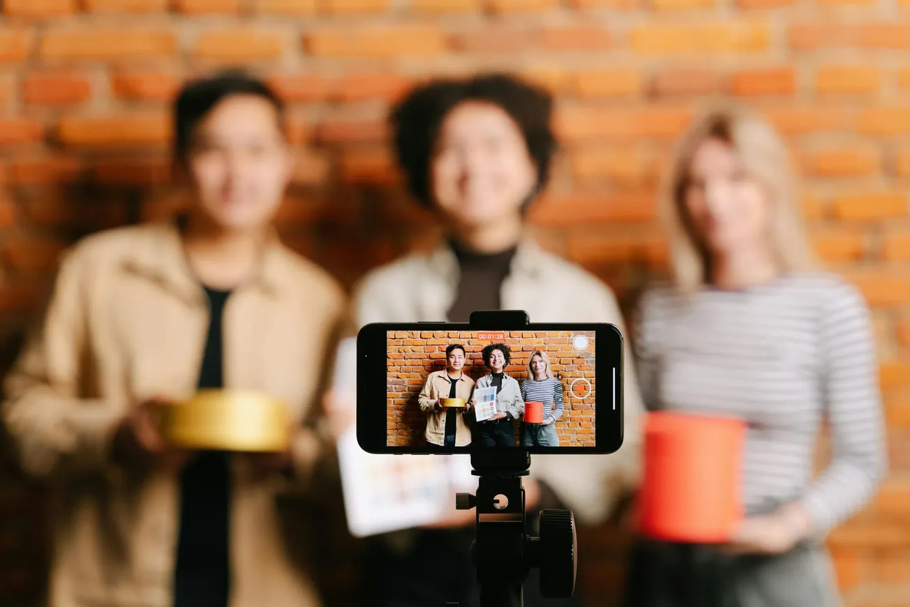A smartphone on a tripod records three blurry people holding objects against a brick wall. The phone display shows them clearly smiling and posing.