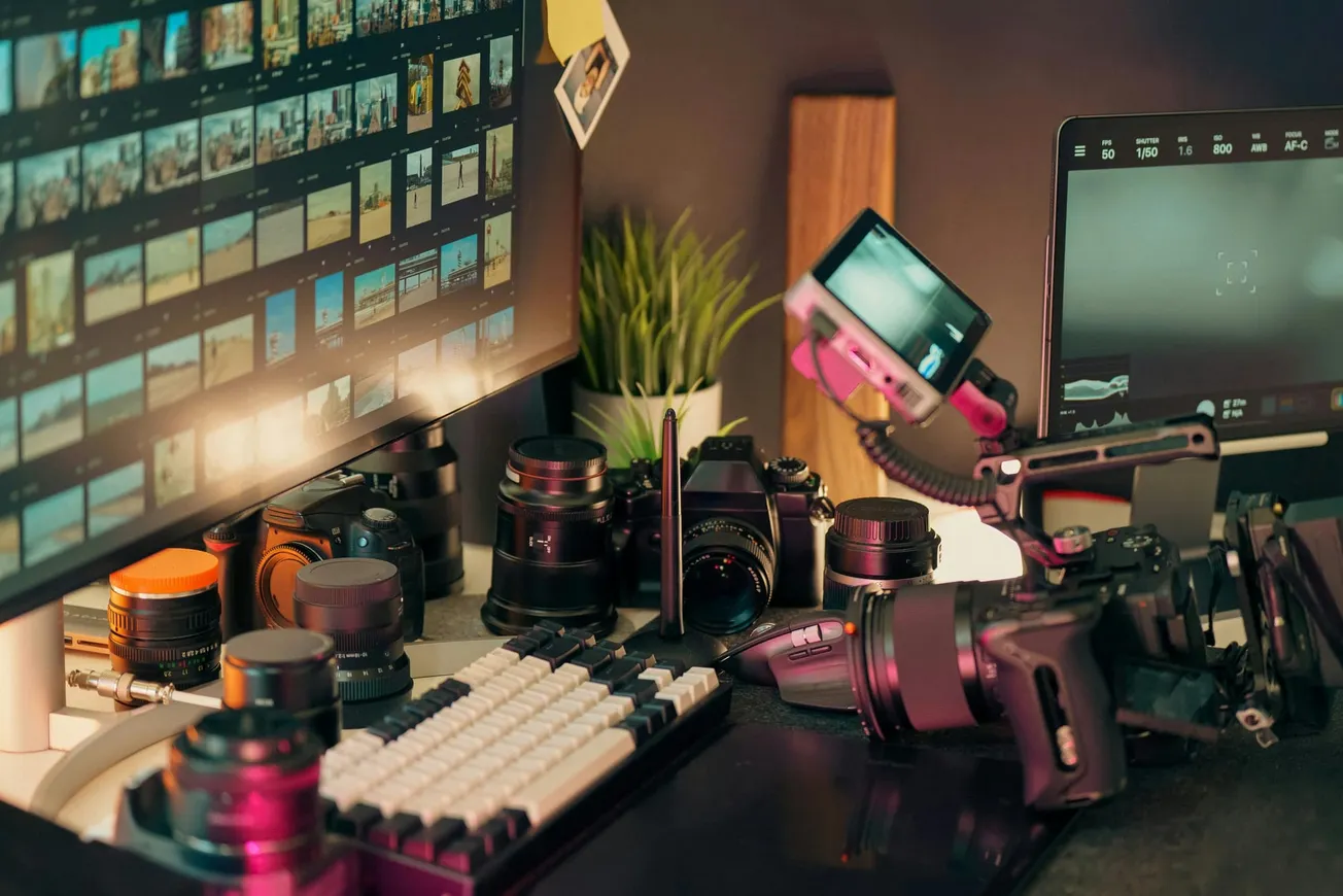 A photographer's workspace with cameras, lenses, a keyboard, and monitor displaying photo thumbnails.