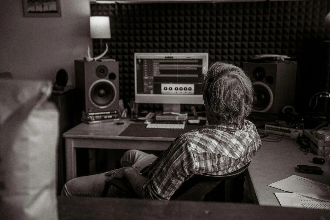 A man in a plaid shirt sits in a dimly lit studio, facing a computer screen with audio editing software. Large speakers and acoustic panels surround him.