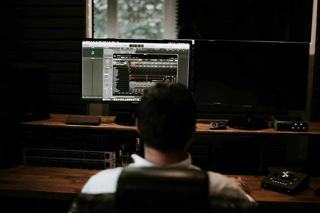 A person sits in a dimly lit recording studio, focusing on two large computer monitors. Audio editing software is open on one screen, creating a focused, creative atmosphere.