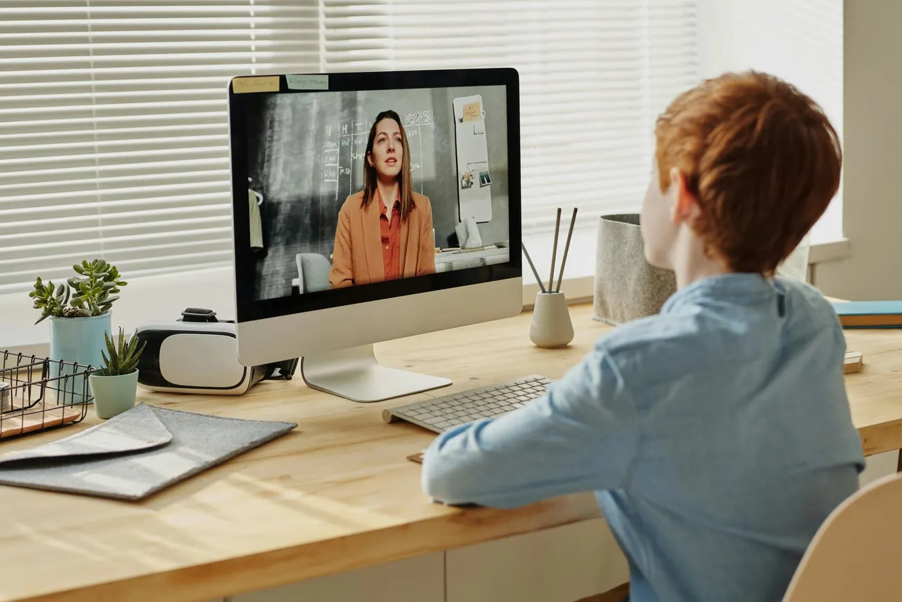 A child with red hair, seated at a wooden desk, attentively watches a virtual lesson on a computer.