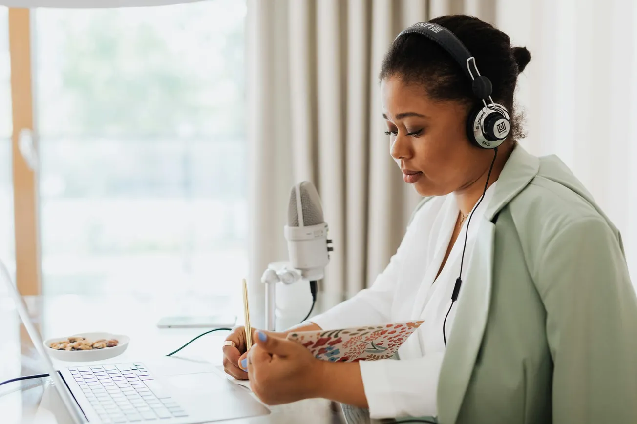A woman wearing headphones sits at a desk with a laptop, microphone, and notepad, focused and serious, creating content or podcasting.
