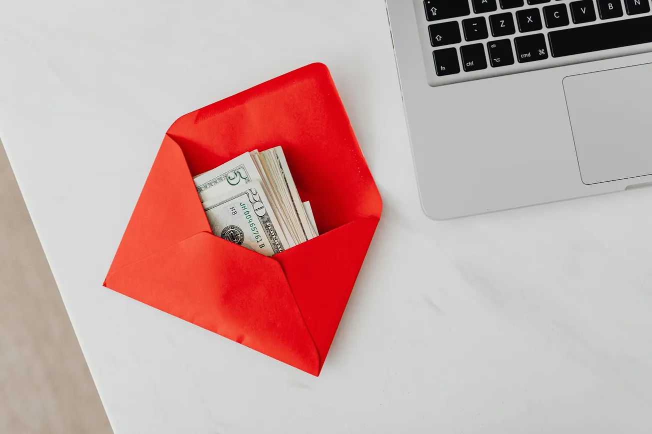 A red envelope containing U.S. dollars is placed on a white table next to a silver laptop. The scene suggests themes of finance or gifting.