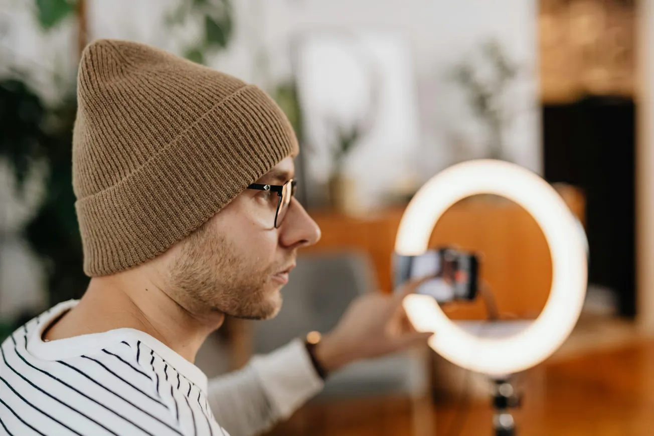 A man wearing a brown beanie and glasses films himself using a smartphone. He is in front of a glowing ring light, conveying a focused, creative atmosphere.