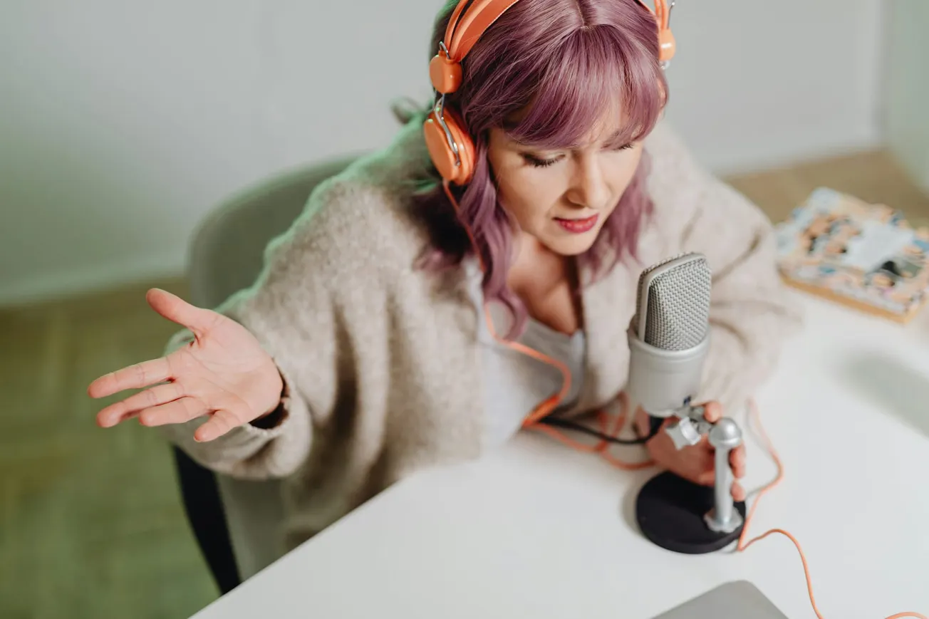 Woman with purple hair and orange headphones recording a podcast. She's speaking into a microphone with an expressive gesture. Cozy, creative atmosphere.