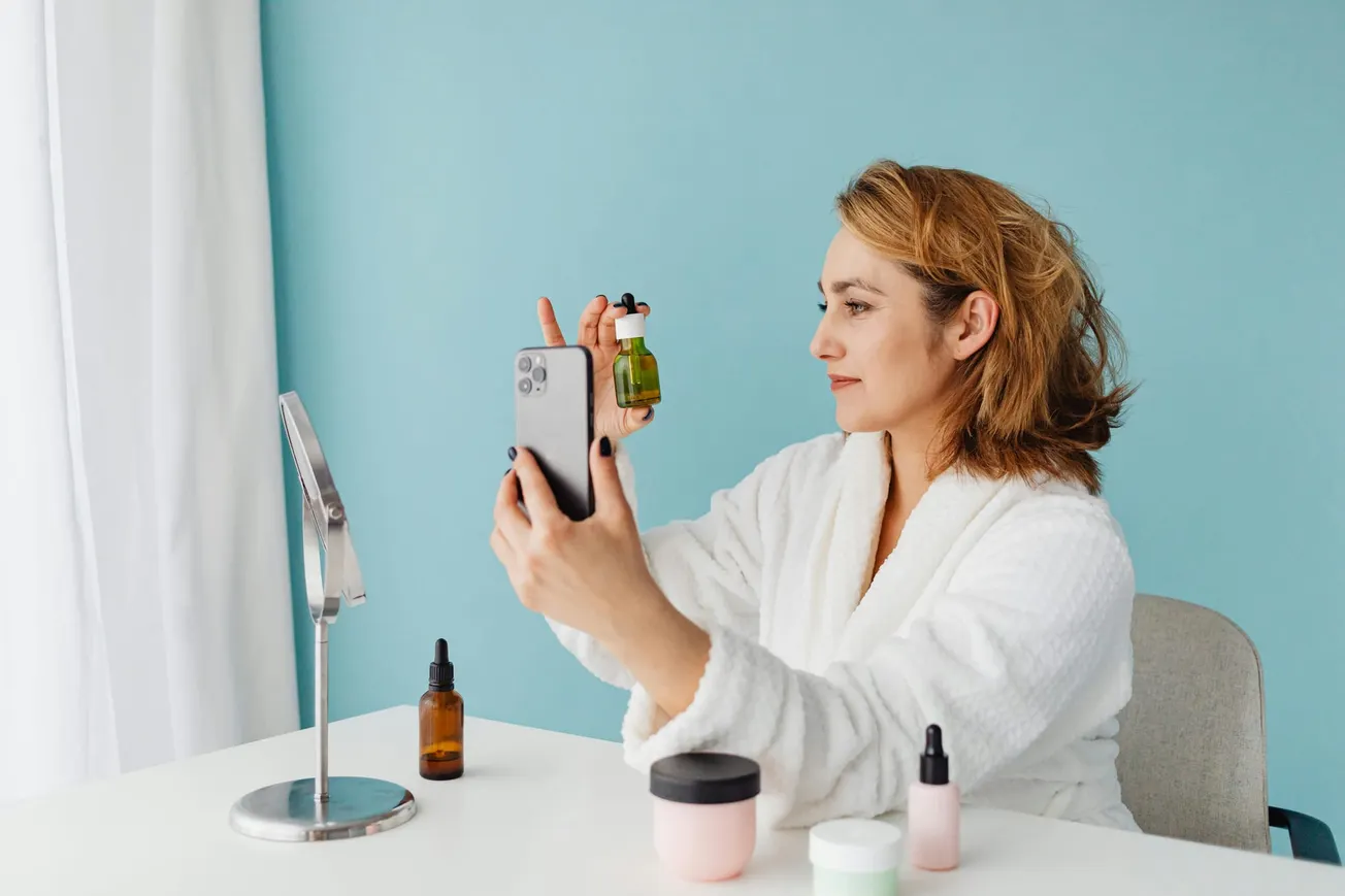 Woman in a white robe holds a green serum bottle and takes a selfie, seated at a vanity with cosmetic bottles. The scene is calm with a light blue background.