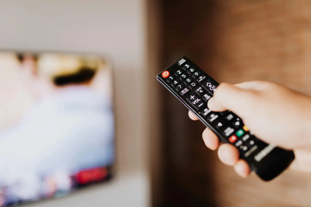 Hand holding a remote control pointed at a blurred TV screen, symbolizing leisure and entertainment.
