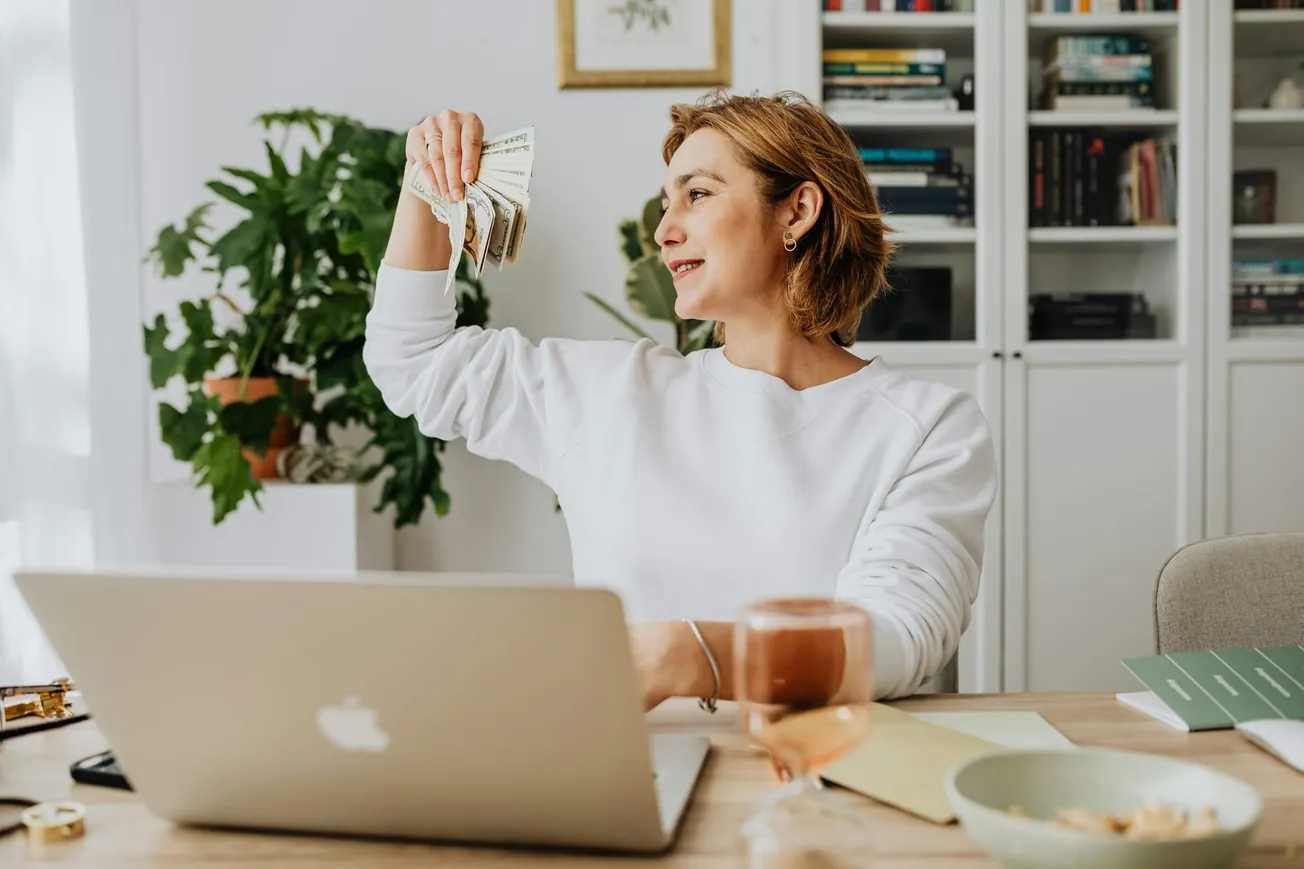 A woman in a white sweater holds a fan of dollar bills, smiling at her laptop. She's sitting at a desk with a glass of wine, exuding a joyful, successful vibe.
