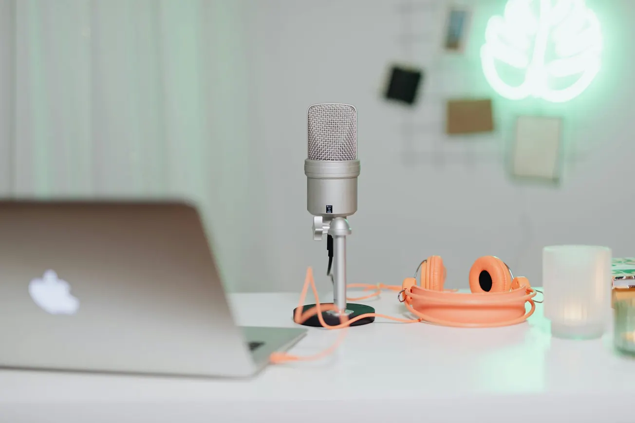 A sleek podcast setup with a gray microphone, orange headphones, and a laptop on a white desk. A soft green neon leaf light glows in the background.