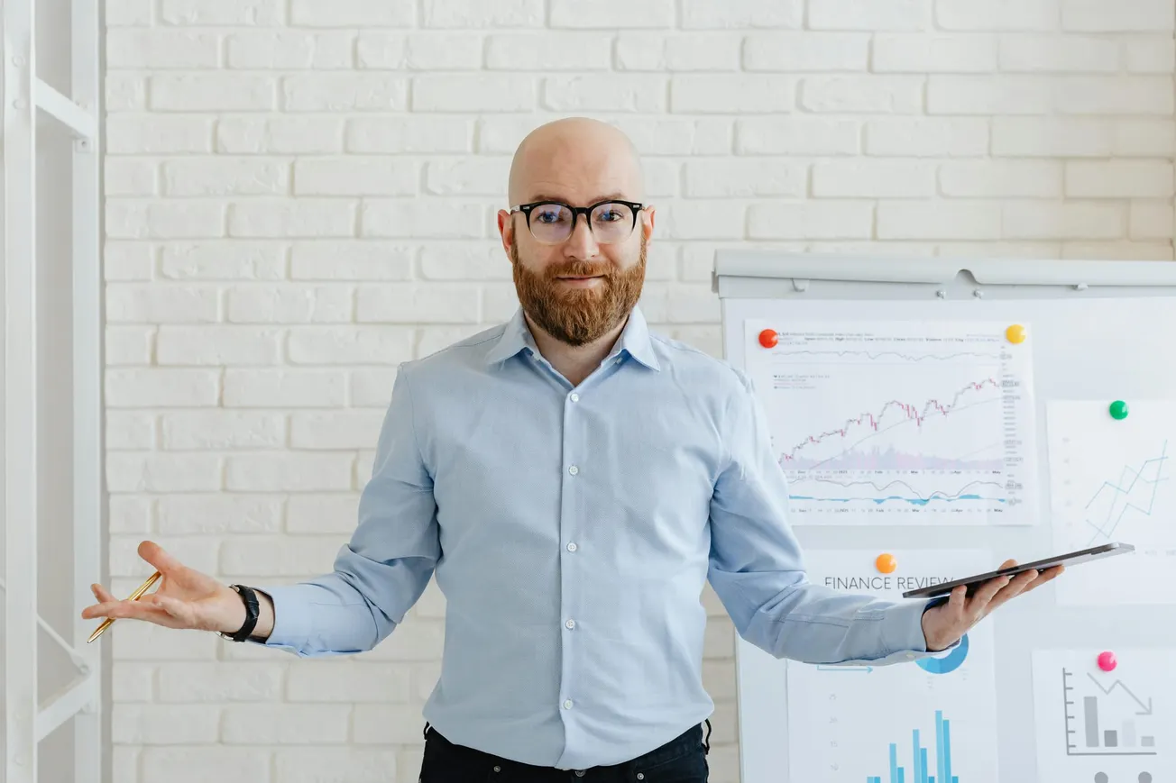 Bald man with a beard wearing glasses and a blue shirt stands confidently holding a tablet. He gestures in front of a chart-filled whiteboard.