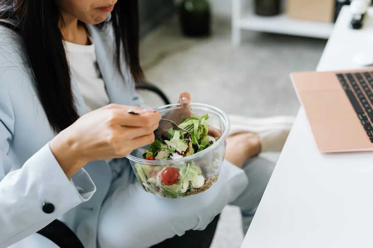 A person wearing a light blue suit sits at a desk, eating a fresh salad from a clear bowl while working on a laptop.