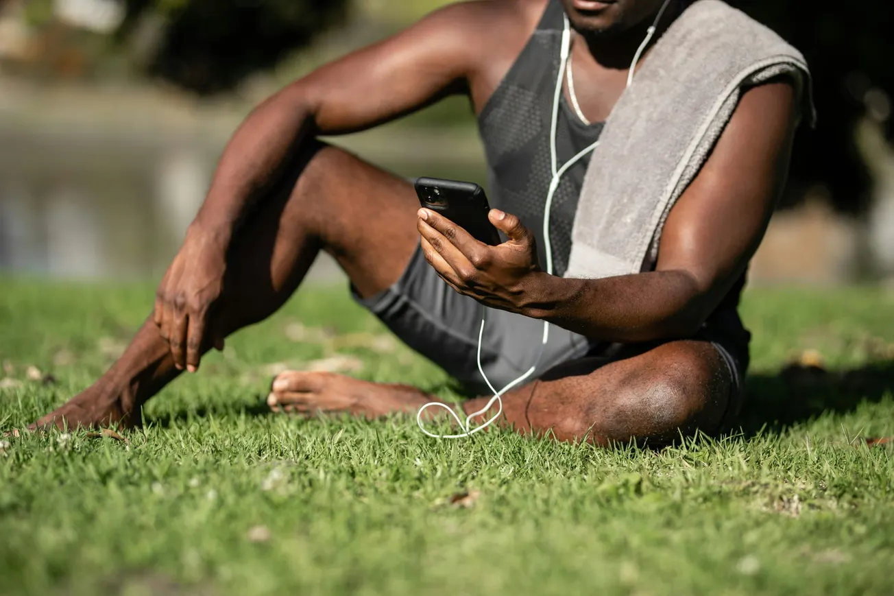 A man sits on grass with a towel on his shoulder, checking his phone. He's wearing workout gear and earphones, suggesting relaxation after exercise.