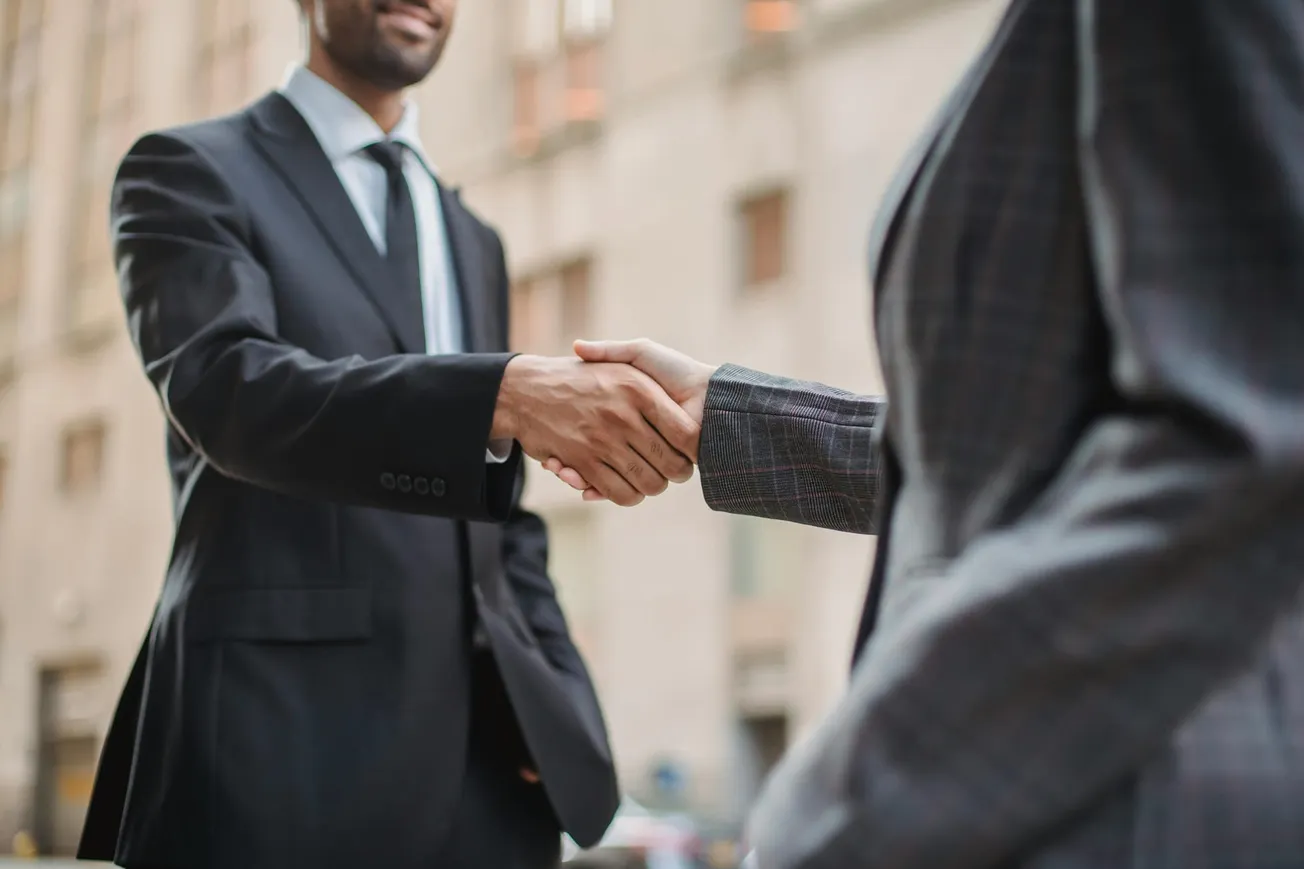 Two people in suits shake hands outdoors, suggesting a business agreement or partnership. The background is a blurred cityscape.