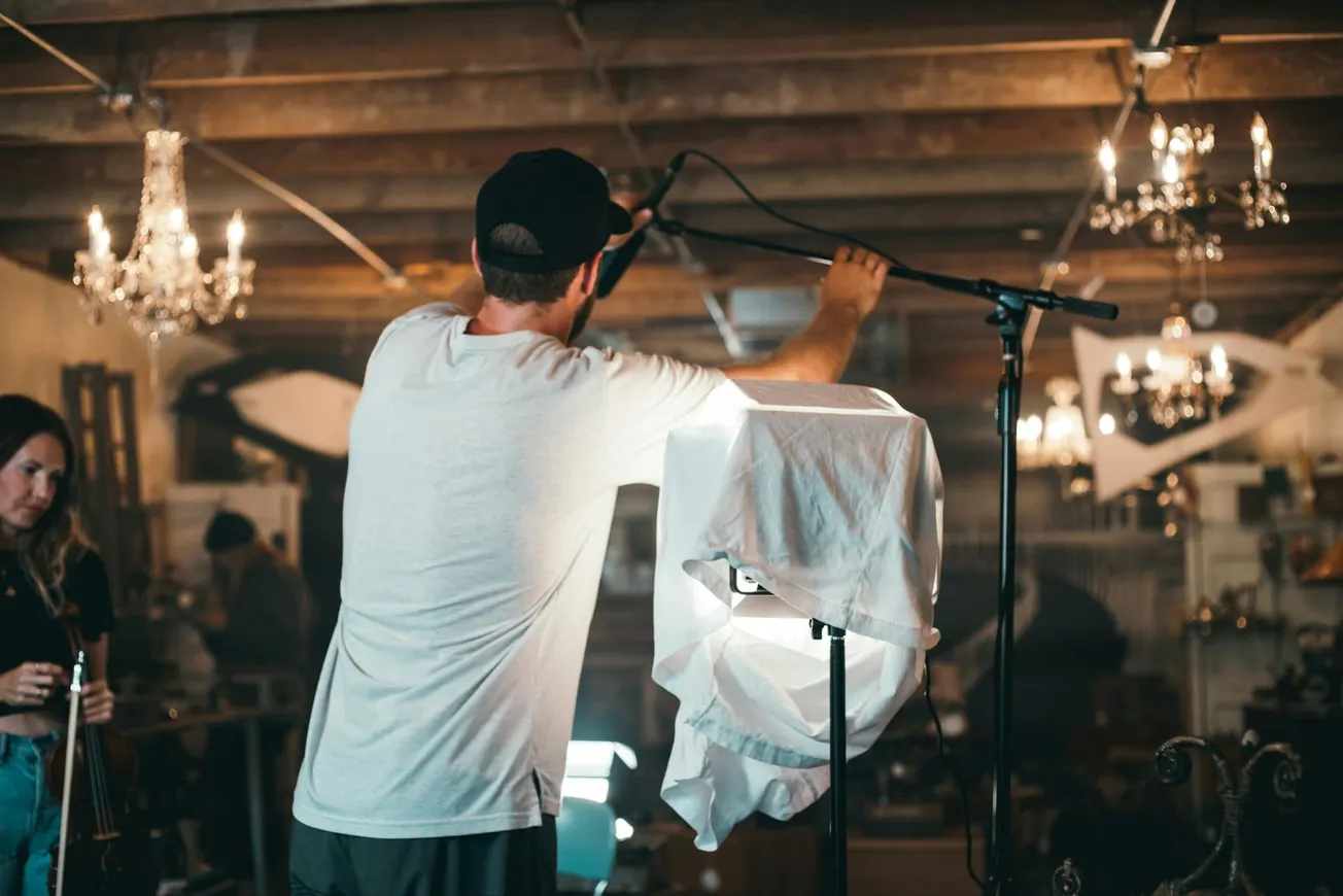 A man in a cap adjusts a microphone in a rustic studio with exposed beams and chandeliers. A covered light stand and focused atmosphere suggests a creative setting.