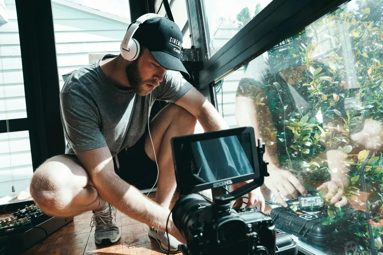 A man crouches indoors, adjusting audio equipment near a large window, wearing headphones and a black cap. A camera with a screen is in the foreground.