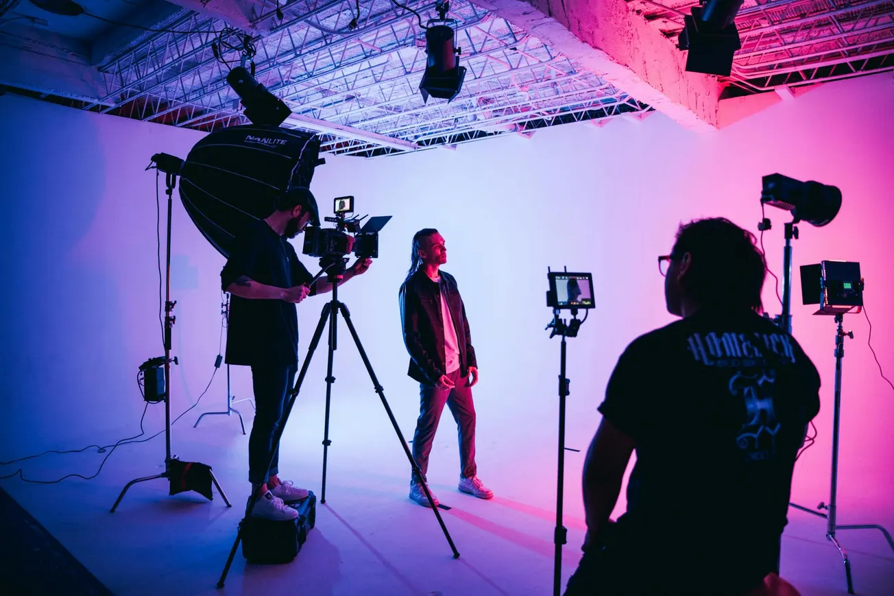 A photoshoot in a studio with pink and purple lighting. A subject stands in the center, while two crew members adjust cameras and equipment, creating a creative and focused atmosphere.
