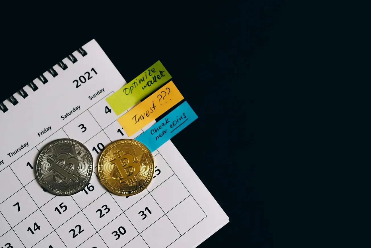 Close-up of a 2021 calendar with silver and gold Bitcoin coins on it, alongside sticky notes reading "Optimize wallet," "Invest???," and "Check new coins."