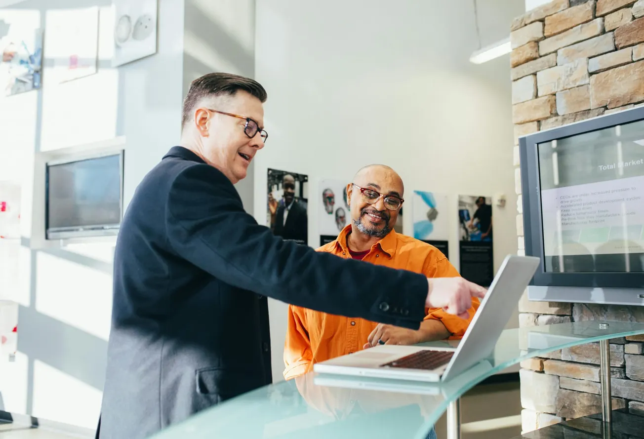 Two men stand at a laptop in a bright office. One, in a suit, points at the screen while smiling. The other, in an orange shirt, looks on, appearing pleased.