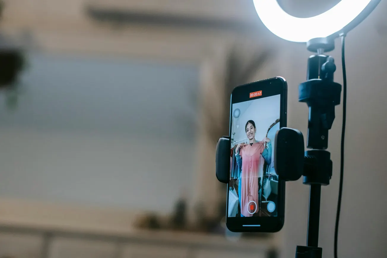 Smartphone on a tripod with a ring light shows a person in a pink dress, smiling during a video recording. The setting is cheerful and creative.