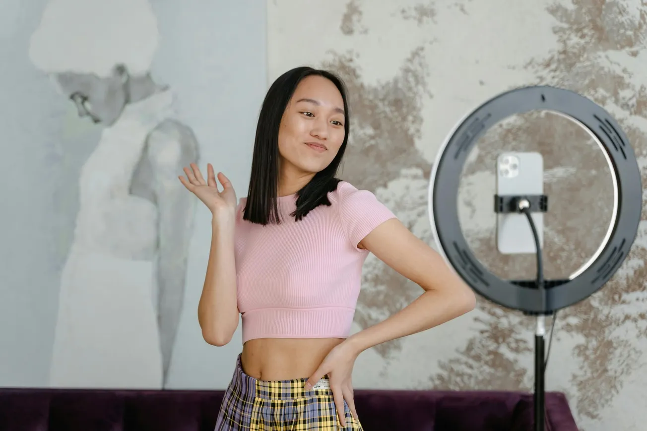 A woman with long hair poses confidently in front of a smartphone held by a ring light stand. She wears a pink crop top and plaid skirt.