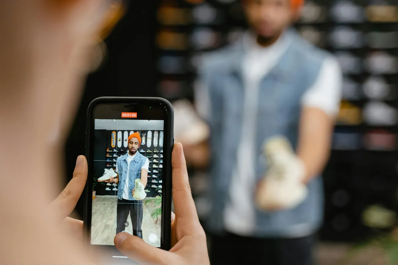 A person records another holding sneakers on a smartphone. The scene is in a shoe store with sneakers lining the wall.