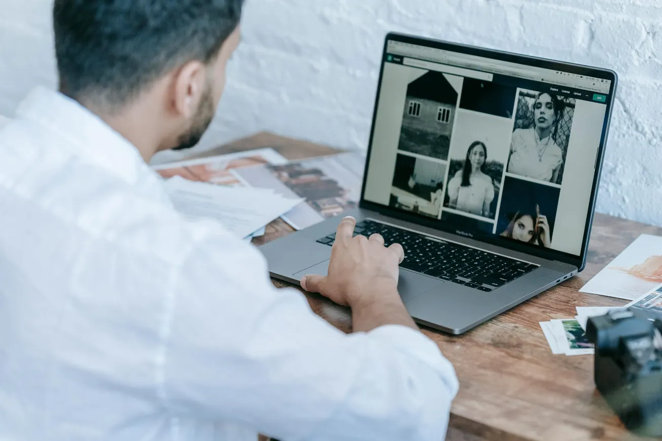 A man in a white shirt is sitting at a wooden table, browsing black and white photos on a laptop. Nearby, papers and a camera are scattered. The mood is focused.