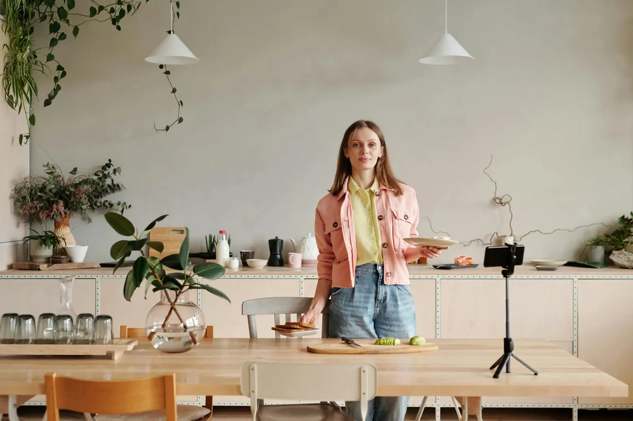 A woman in a pink jacket and blue jeans stands in a modern kitchen, holding plates. The table has plants, glassware, and a smartphone on a tripod.