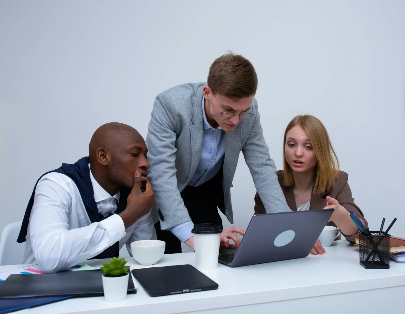 Three people engaged in discussion around a laptop in an office setting. They appear focused and collaborative, with notebooks and coffee on the table.