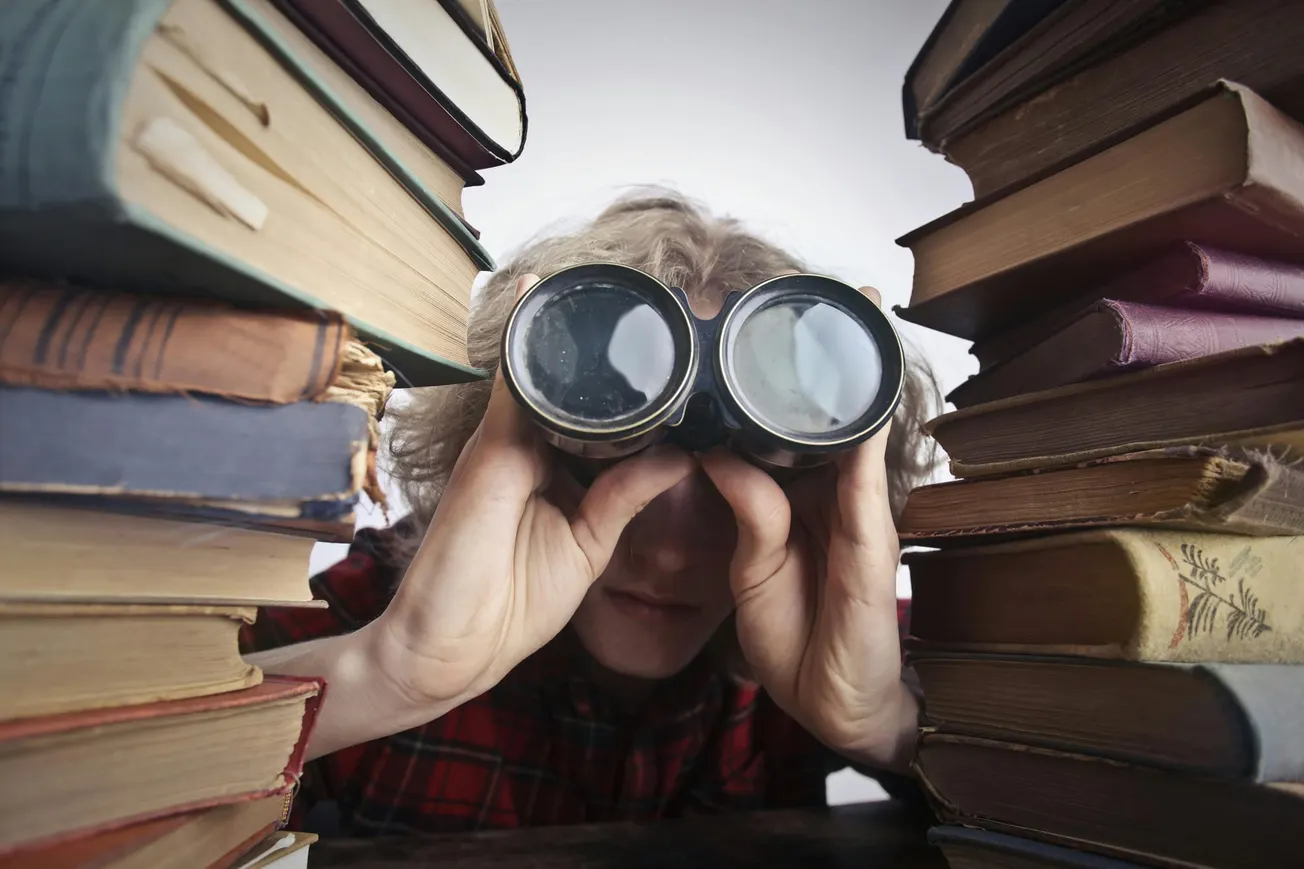 A person peers through binoculars, surrounded by stacks of old books. The setting conveys curiosity and a sense of exploration or research.