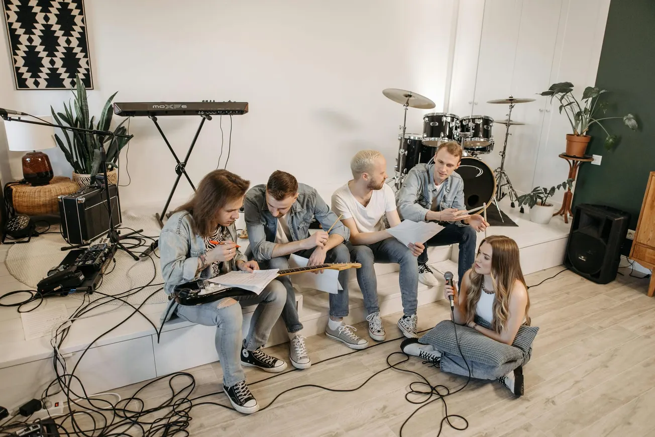 A group of five people seated in a music studio, discussing sheet music. Instruments like a guitar, keyboard, and drums are visible.