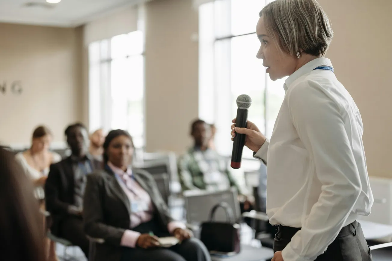 A person in a white shirt is speaking into a microphone, addressing an attentive audience in a conference room.