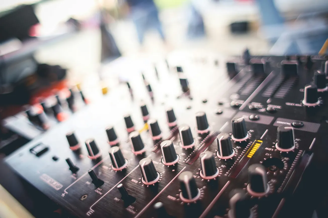 Close-up of a DJ mixer with numerous knobs and sliders, softly lit, creating an energetic atmosphere. The blurred background suggests a lively setting.