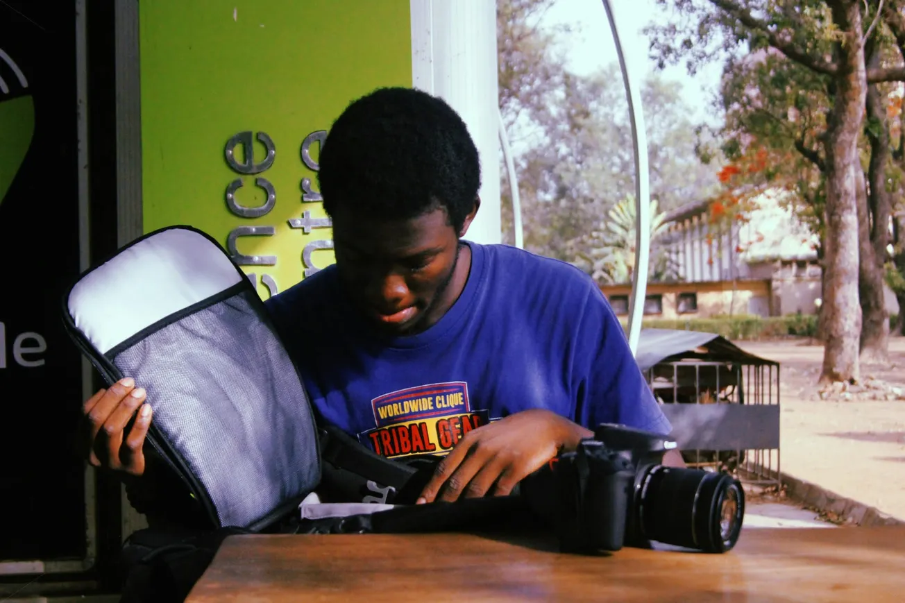 A person in a blue t-shirt looks into an open camera bag on a wooden table, with a camera nearby. Green signage and trees are in the background.
