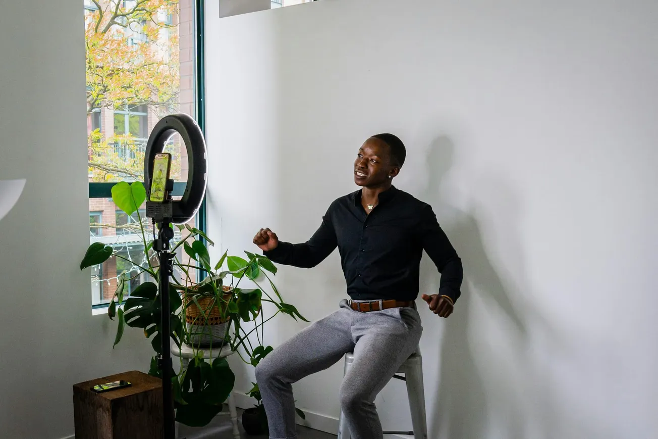 A person in a black shirt and gray pants sits on a stool facing a ring light with a smartphone. They appear joyful, with plants and a window behind.