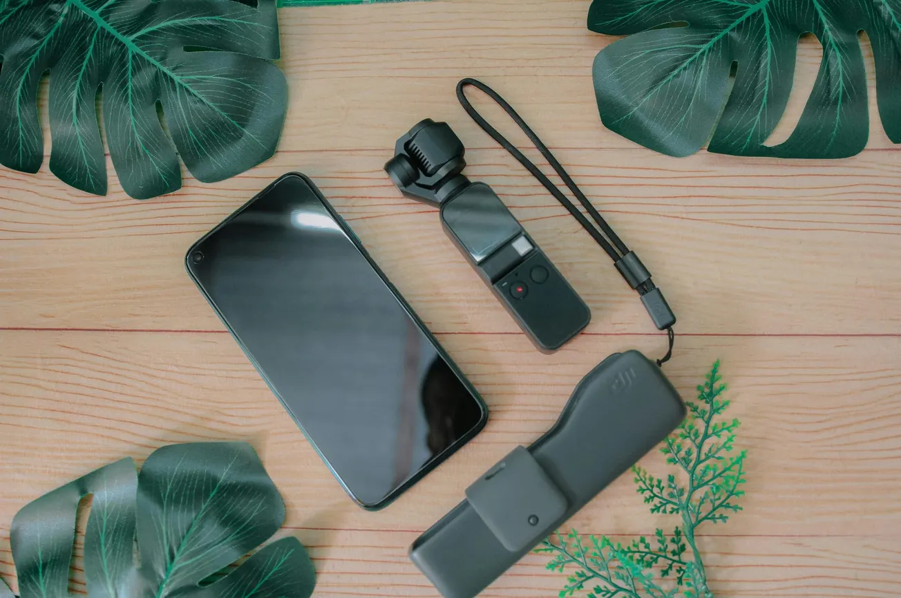 Smartphone, compact camera, and case on a light wooden surface surrounded by green tropical leaves.