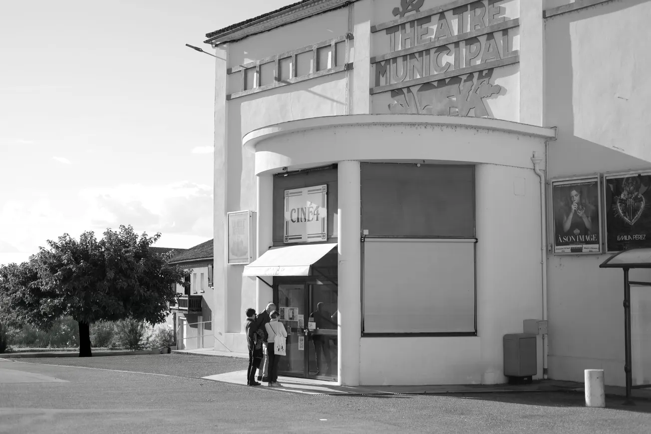 Black-and-white photo of a municipal theater entrance with two people checking posters. The tone is serene, with a tree and clear sky in the background.