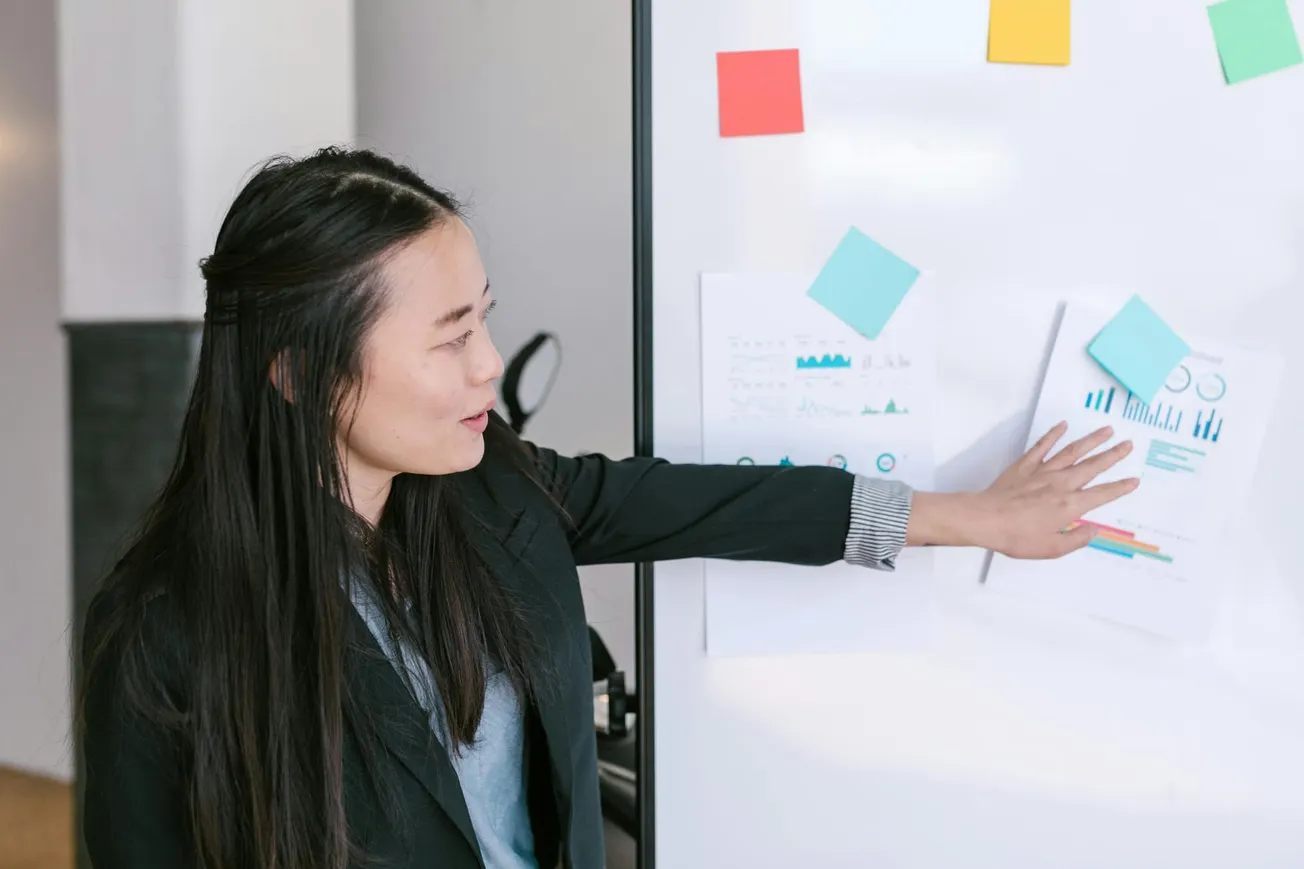 A woman in a black blazer points to graphs on a whiteboard adorned with colorful sticky notes.