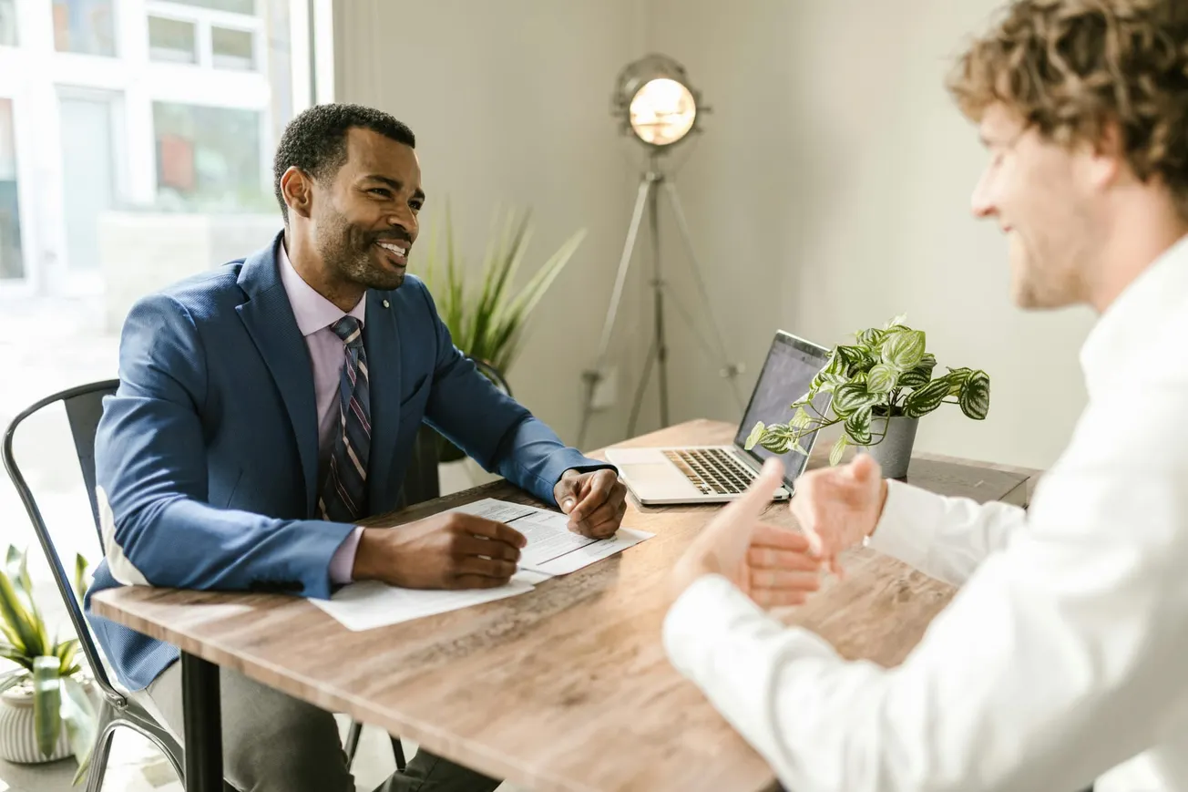 Two men talk at a desk. One wears a blue suit and smiles, while the other gestures with hands. A laptop and plant are on the table.