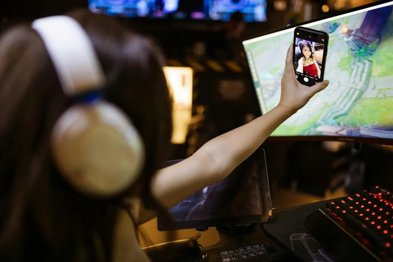 Person with headphones takes a selfie while sitting at a gaming setup. They're holding a smartphone, with a computer screen displaying a game.