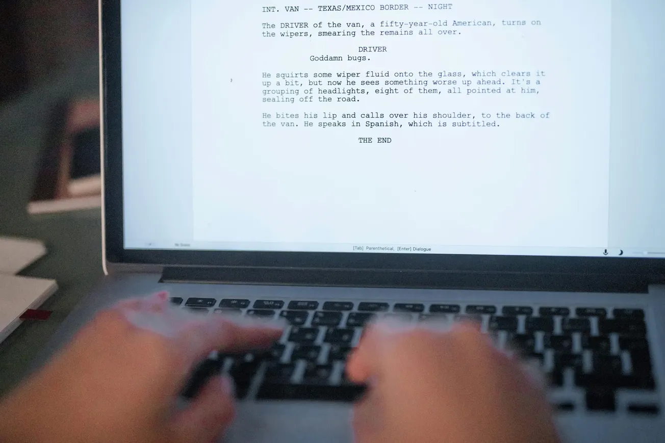 Blurred hands type on a laptop keyboard, focusing on a screenplay displayed on the screen. The script mentions a driver at the Texas/Mexico border.