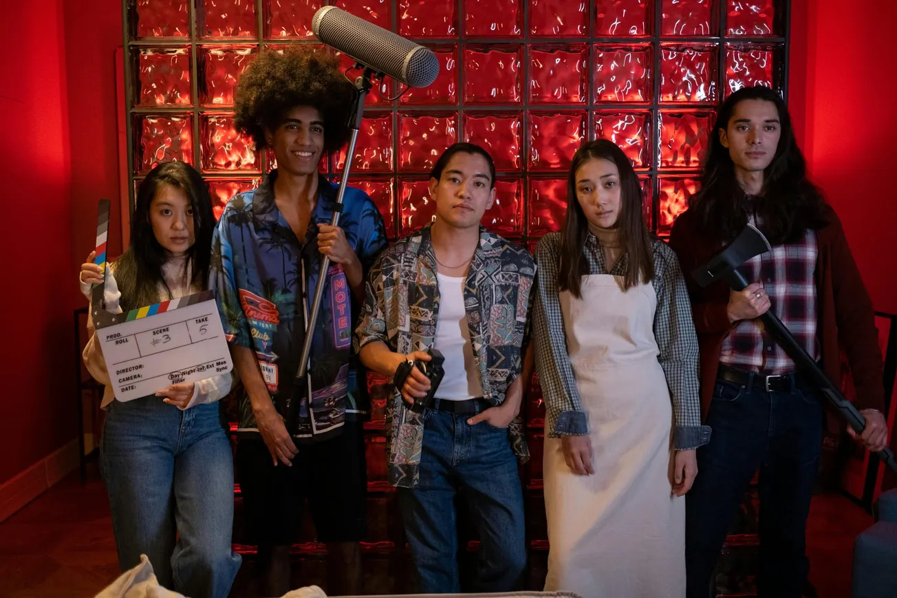 A diverse group of five people stand in front of a red glass block wall, holding film equipment, including a clapperboard and microphone, in a cinematic setting.