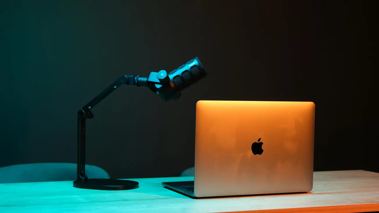A podcast setup with a microphone on an adjustable arm and a laptop on a wooden desk. The ambient lighting is a mix of blue and orange.