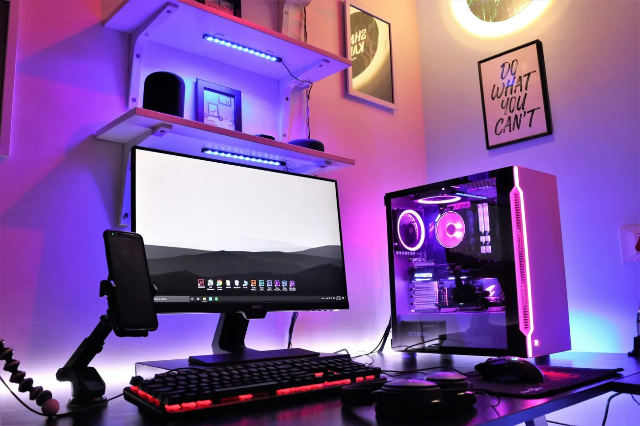 A modern gaming setup featuring a desktop PC with vibrant purple LED lights, a sleek monitor, keyboard, and mouse. Shelves hold accessories, and motivational posters adorn the wall.