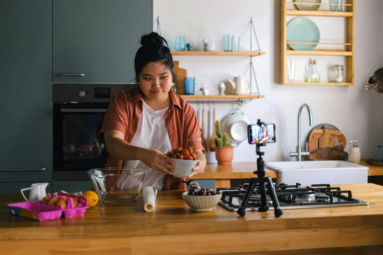 A person in a kitchen holds a bowl of tomatoes, preparing a meal on a counter with ingredients and a smartphone on a tripod. The scene feels focused and casual.