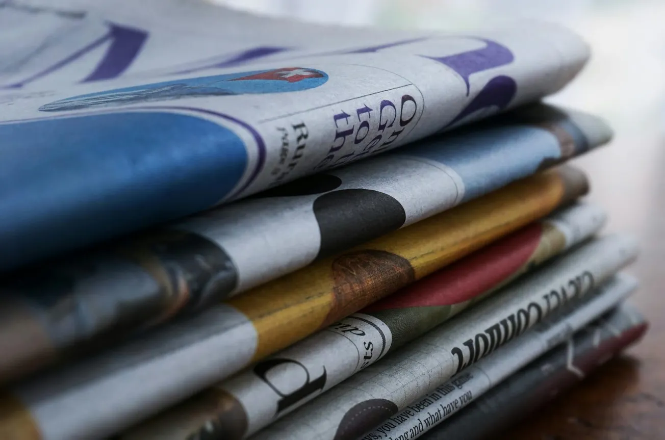 A close-up image of a stack of folded newspapers in various colors. The layered papers create a textured look, conveying a sense of news and information.