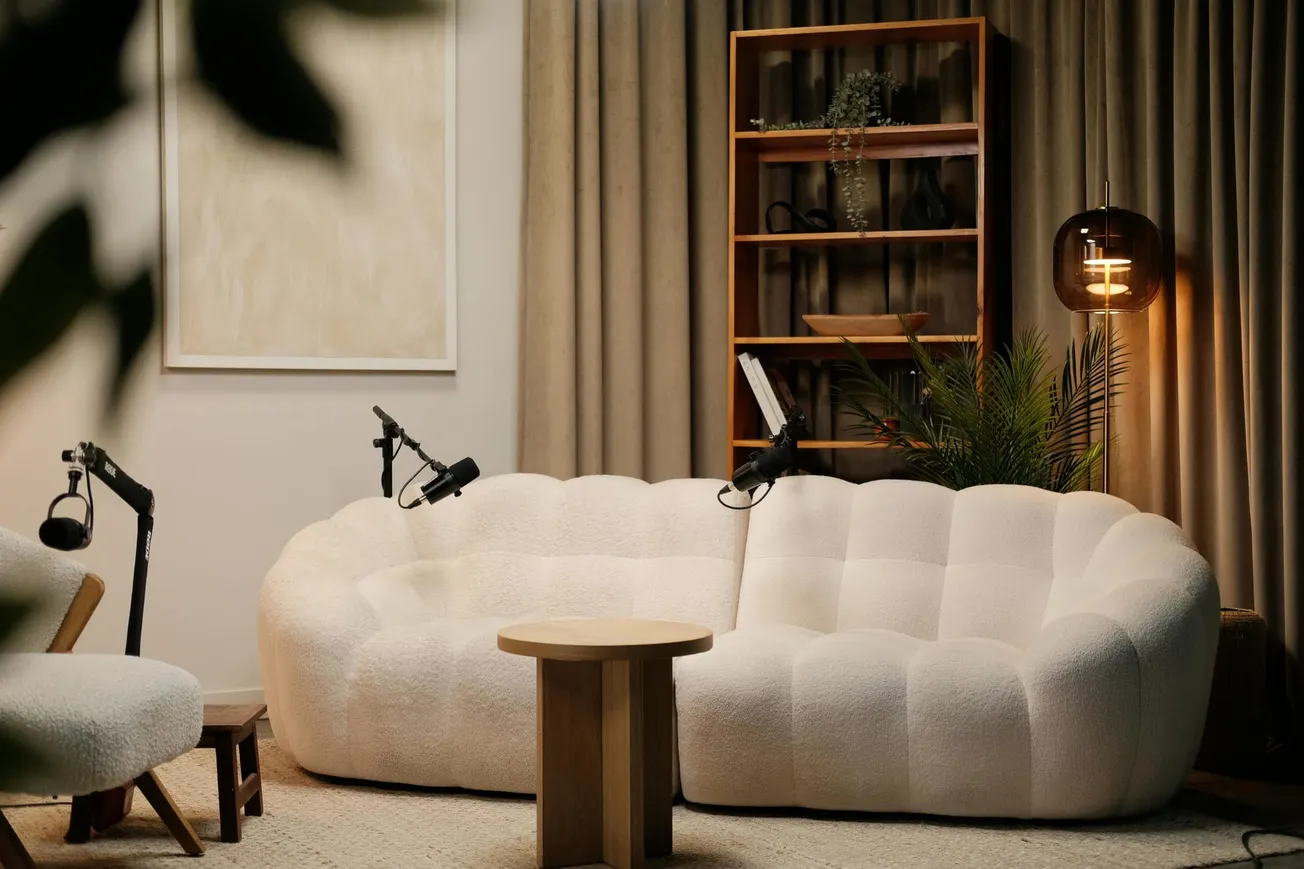 Cozy studio setup with a plush white sofa, wooden table, and microphones. Shelves and plants in the background.