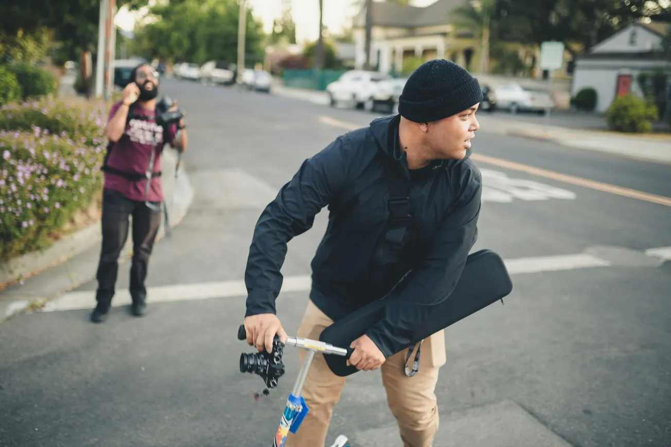 A man in a black jacket and beanie rides a bicycle on a suburban street, carrying a camera. Another person holding a camera in the background appears to be calling out.
