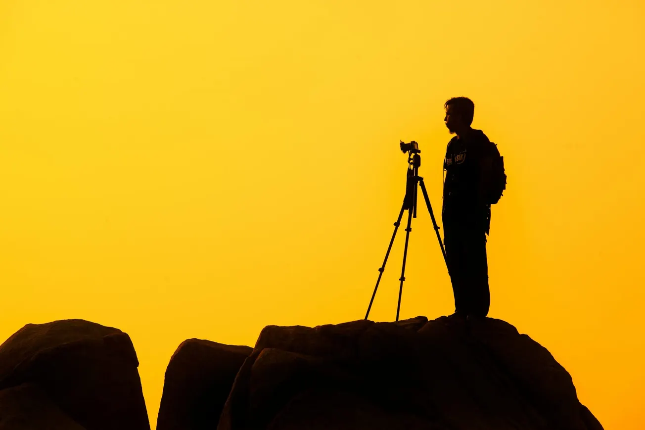 Silhouette of a person with a backpack standing beside a camera on a tripod atop a rocky hill, set against a vibrant orange sunset sky.