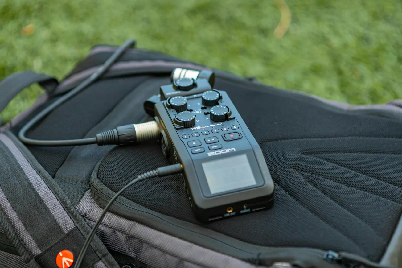 A portable audio recorder placed on a black backpack, with cables connected, lies on a grassy surface, conveying a sense of outdoor recording setup.