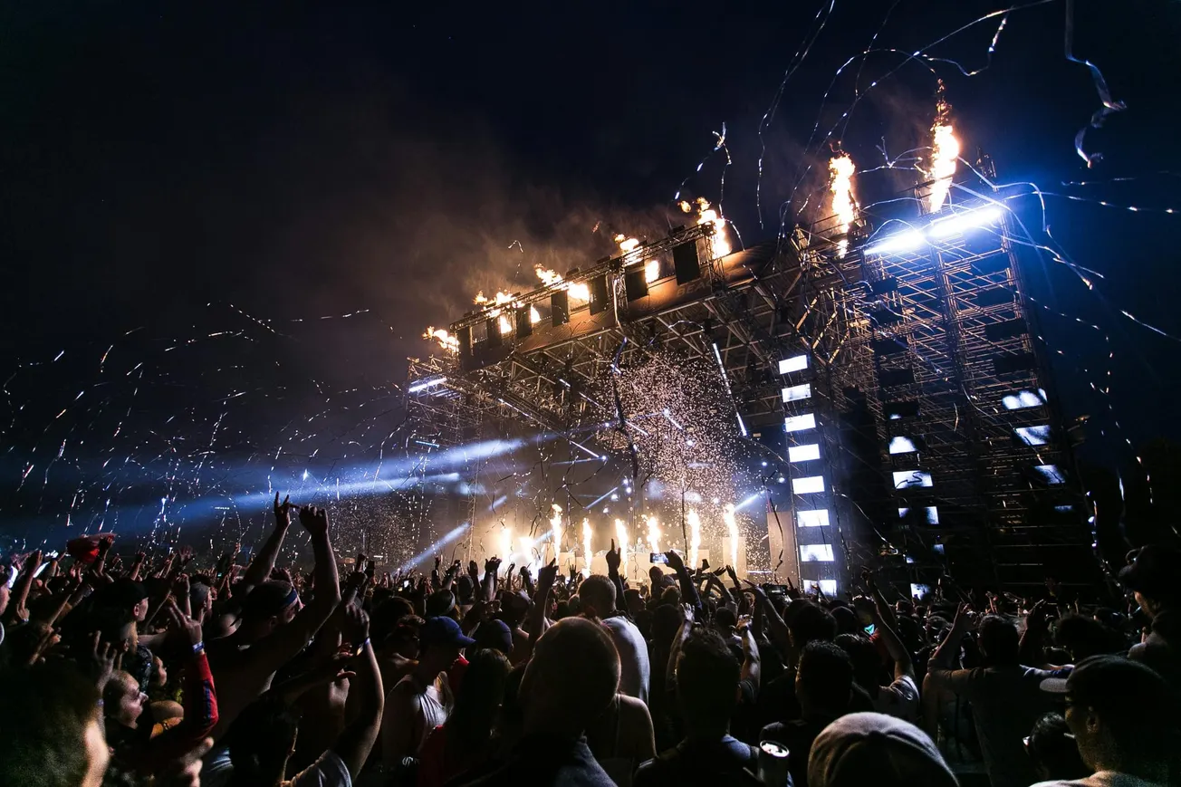 Massive outdoor concert at night with crowd cheering, hands raised. Stage lit with bright lights, fireworks, and confetti, creating an energetic vibe.